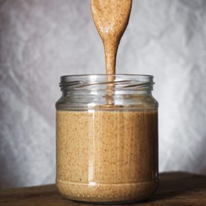 How to make Homemade Almond Butter couldn't be easier, healthier and cheaper! Meade with just almonds, it's smooth and creamy. Shot of a jar with almond butter and a spoon pouring almond butter in it. It can be seen how creamy and runny it is.