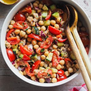 This Mediterranean Chickpea Salad is simple but so flavourful! It combines chickpeas with fresh vegetables and delicious canned tuna, all dressed up with a classic Spanish vinaigrette. It makes a great standalone meal, but it's also perfect as a side or starter. What's more, do you need a packed meal or have a planned picnic or barbecue? This is your salad as it can be prepared in advance and it's so good that everybody will love it. In this overhead shot the salad can be seen served in a bowl with a serving wooden spoon set in it.
