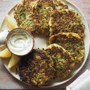 These Easy Courgette Fritters with Lemony Herby Crème Fraîche are a game changer for weeknight meals. They are gluten-free and combine eggs, courgette, and a delicious mix of herbs and spices that make them yummy while being light. In this overhead shot, the fritters can be seen placed on a plate in a semi circular fashion around a little bowl with the crème fraîche dip. A piece of a kitchen cloth can be seen on the bottom right corner.