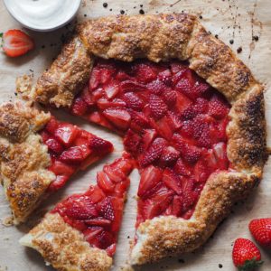 Bursting with fresh spring strawberries this Strawberry Galette with Vanilla Whipped Cream on the side is truly a spring and summer treat. Made with a quick flaky pastry and accompanied with vanilla whipped cream, it's a twist on the classic strawberries and cream dessert that will make everybody WOW. It's delicious, simple and super gorgeous. You can’t go wrong with this one, I promise! Here we can see an overhead shot of the whole galette. 2 portions have been sliced on the left and separated slightly from the rest of the galette. At the top left corner there is a small bowl with the whipped cream with half strawberry on top and half strawberry on its left side. On the bottom right, next to the galette, there are 2 strawberries.